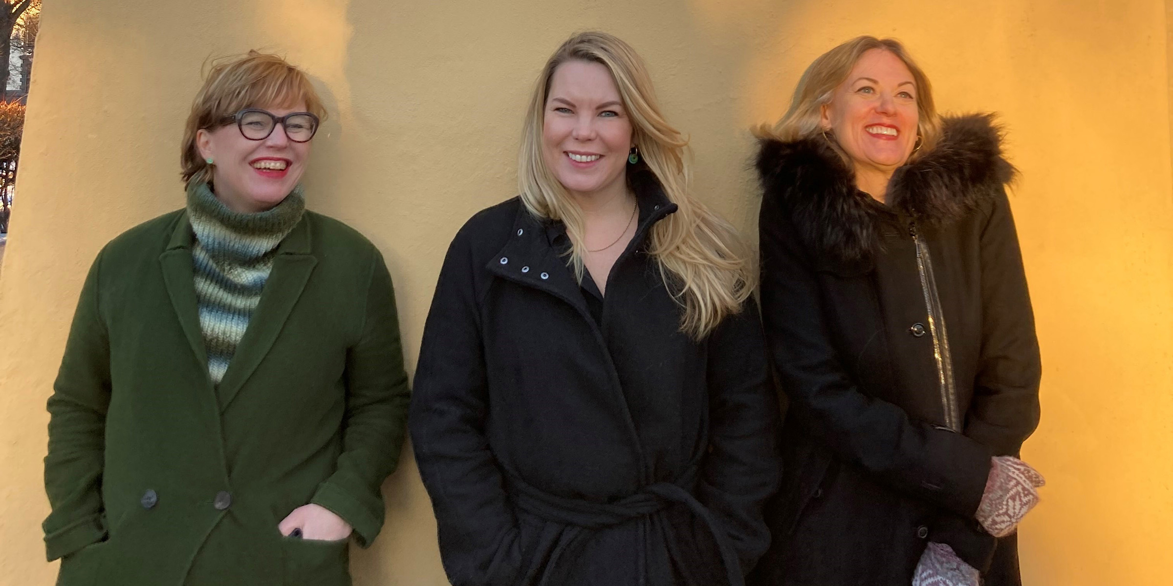 Forfattere Hilde Østby, Marta Breen og Helene Uri står smilende foran en gul murvegg.