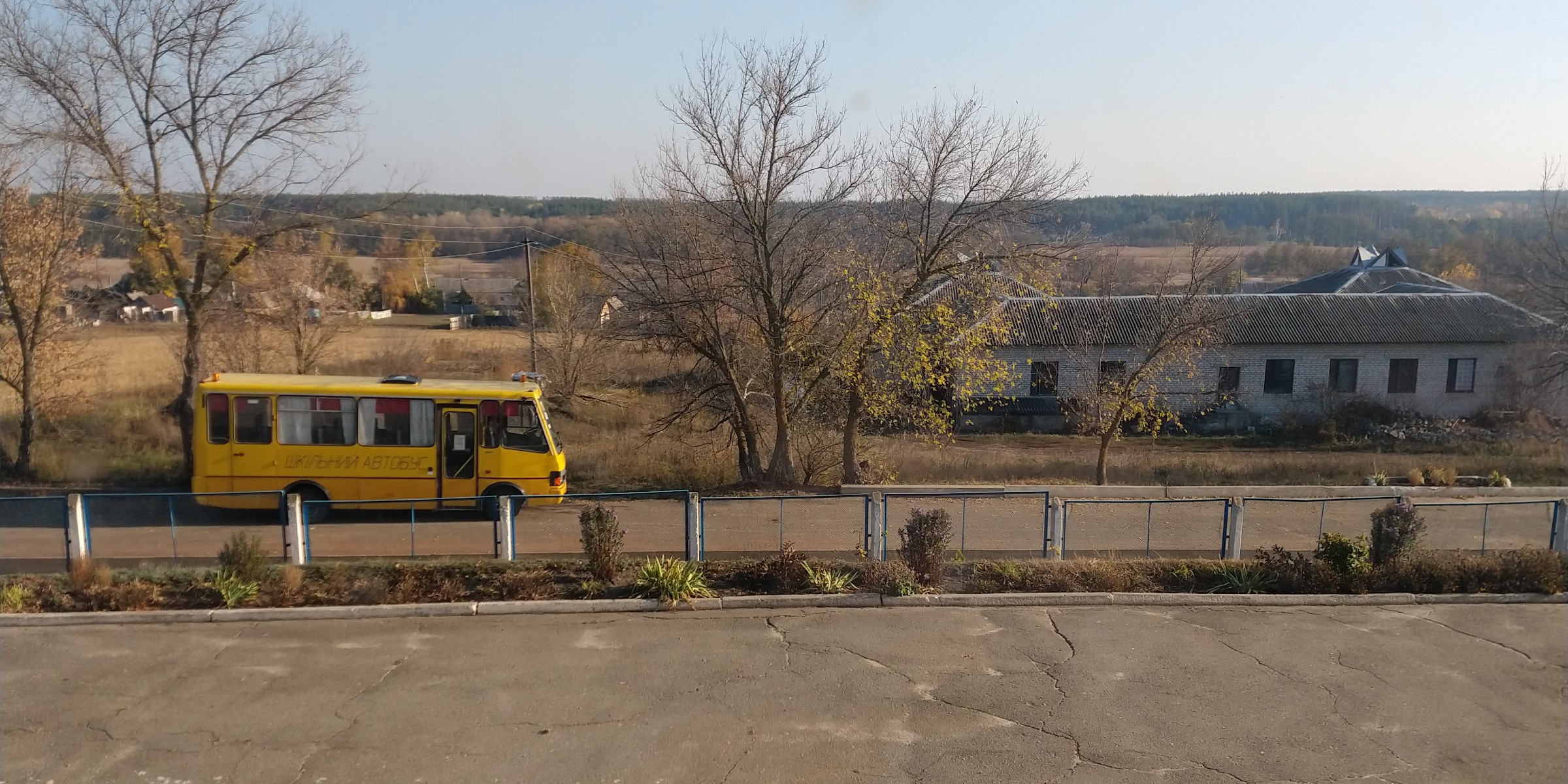 En tom, gul buss står på en asfaltert vei. I bakgrunnen ser vi spredt bebyggelse, åkrer og skog.