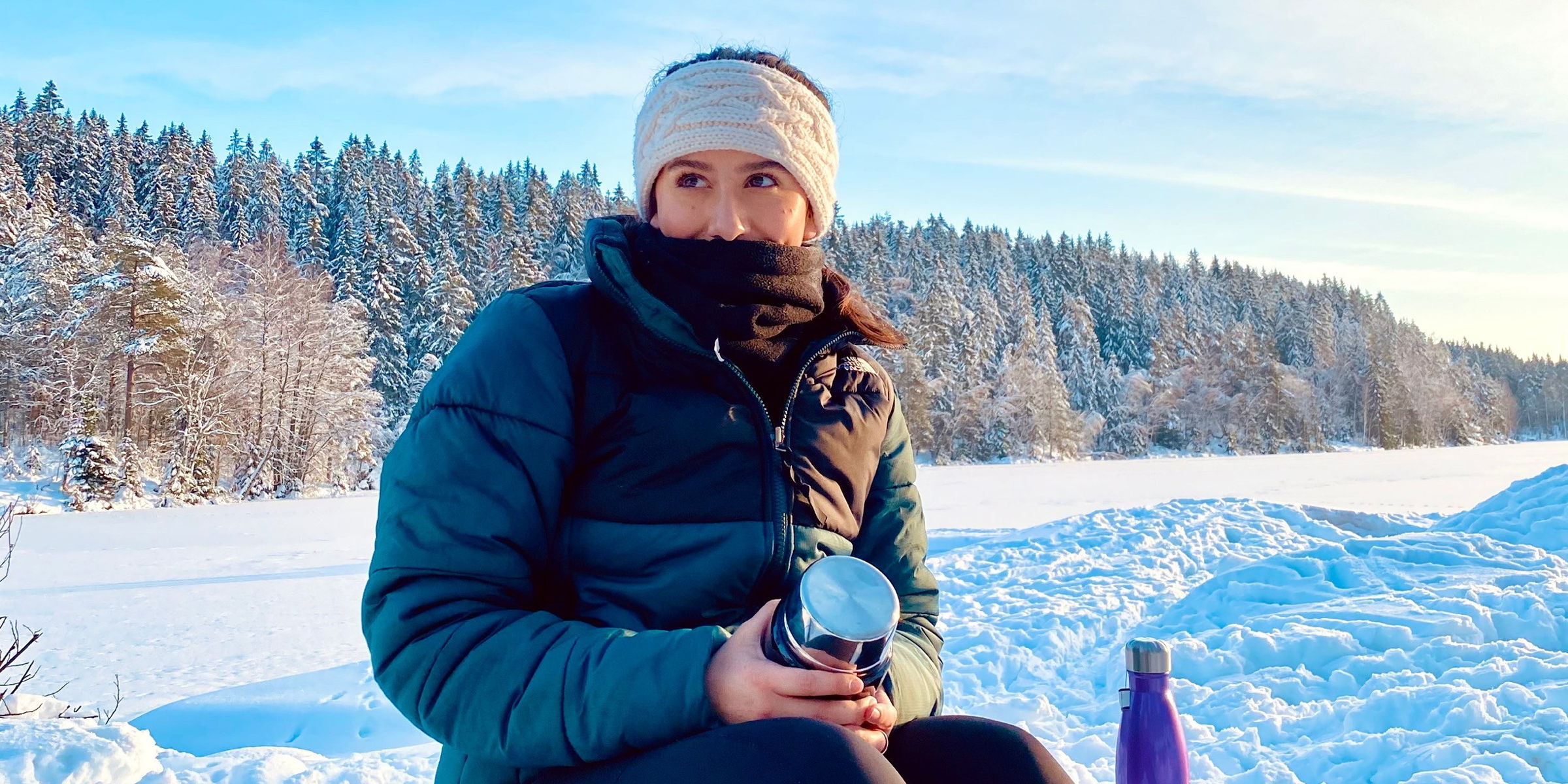 OsloMet student in winter clothes sits on a bench in a snowy landscape. There is forest and blue sky behind her. She is holding a thermos.