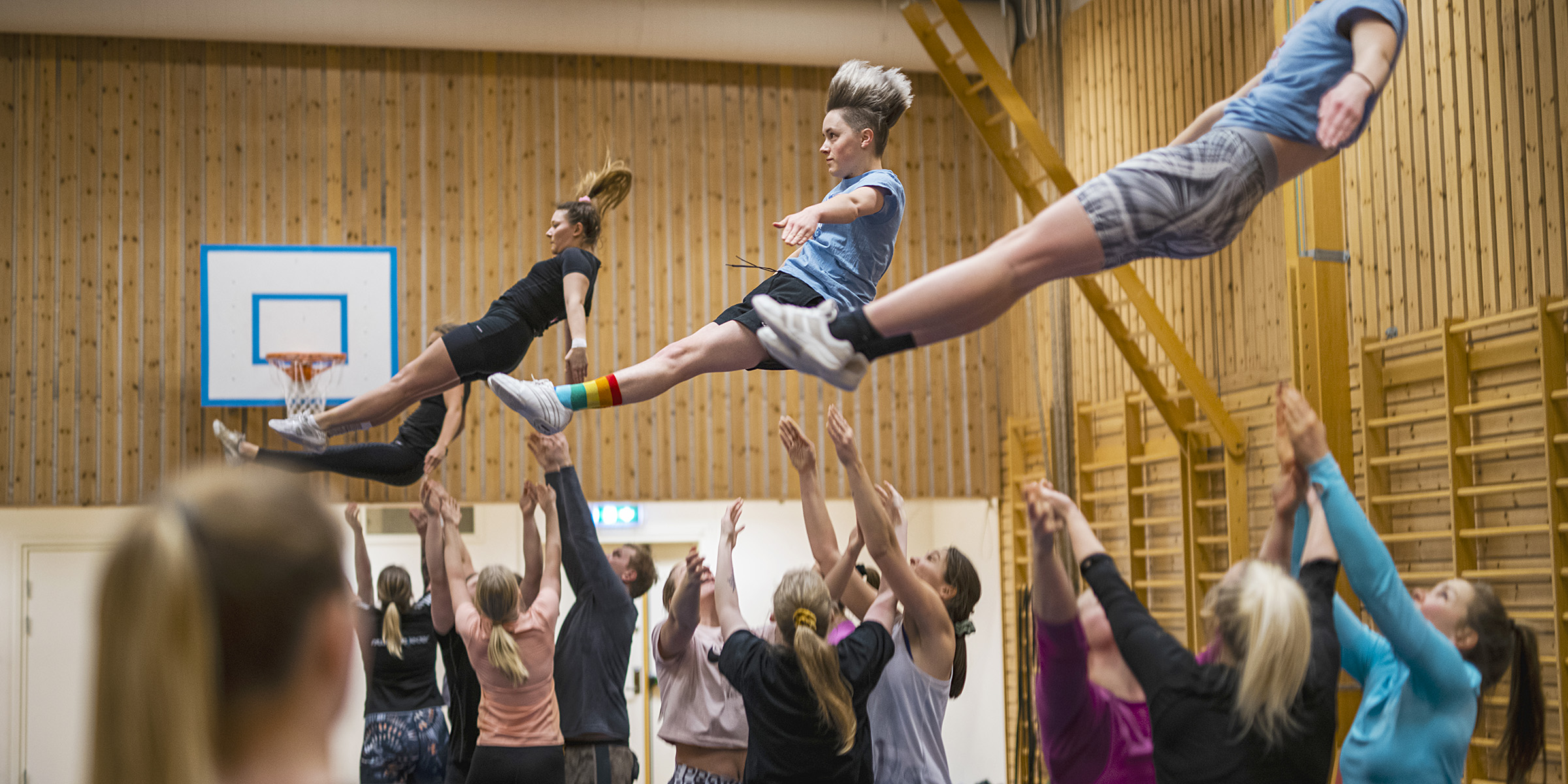 A large group of people practice acrobatics in a gymnasium. Together they throw three people into the air.