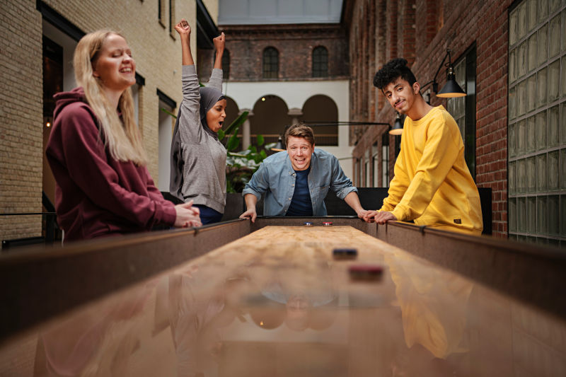 4 studenter som spiller shuffleboard. Ei jente strekker hendene opp i taket, en gutt ser etter pucken han nettopp har sendt avgårde. To studenter står rundt og ser på. 