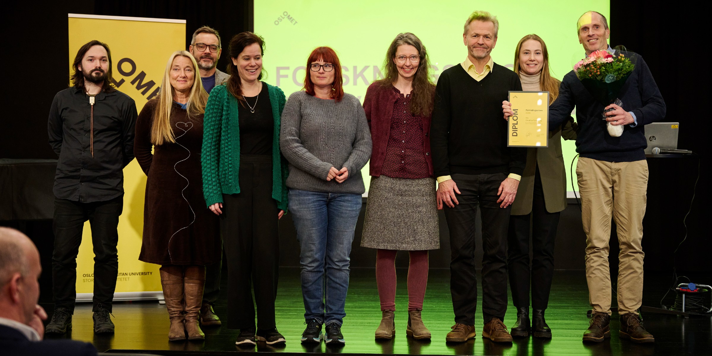 Foto av prisvinnere av formidlingsprisen, stående på scenen med blomst og diplom.