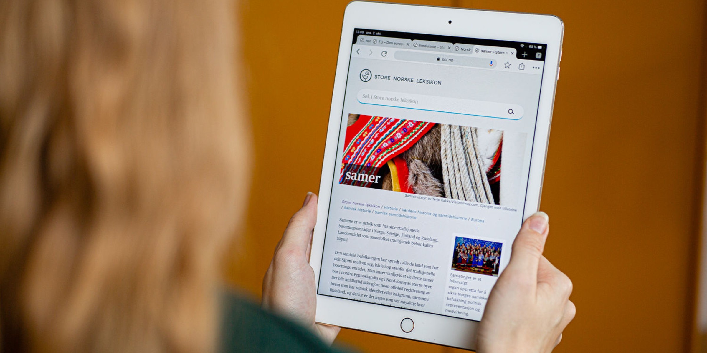 Foto av hender som holder et nettbrett med en artikkel om samer fra Store norske leksikon.