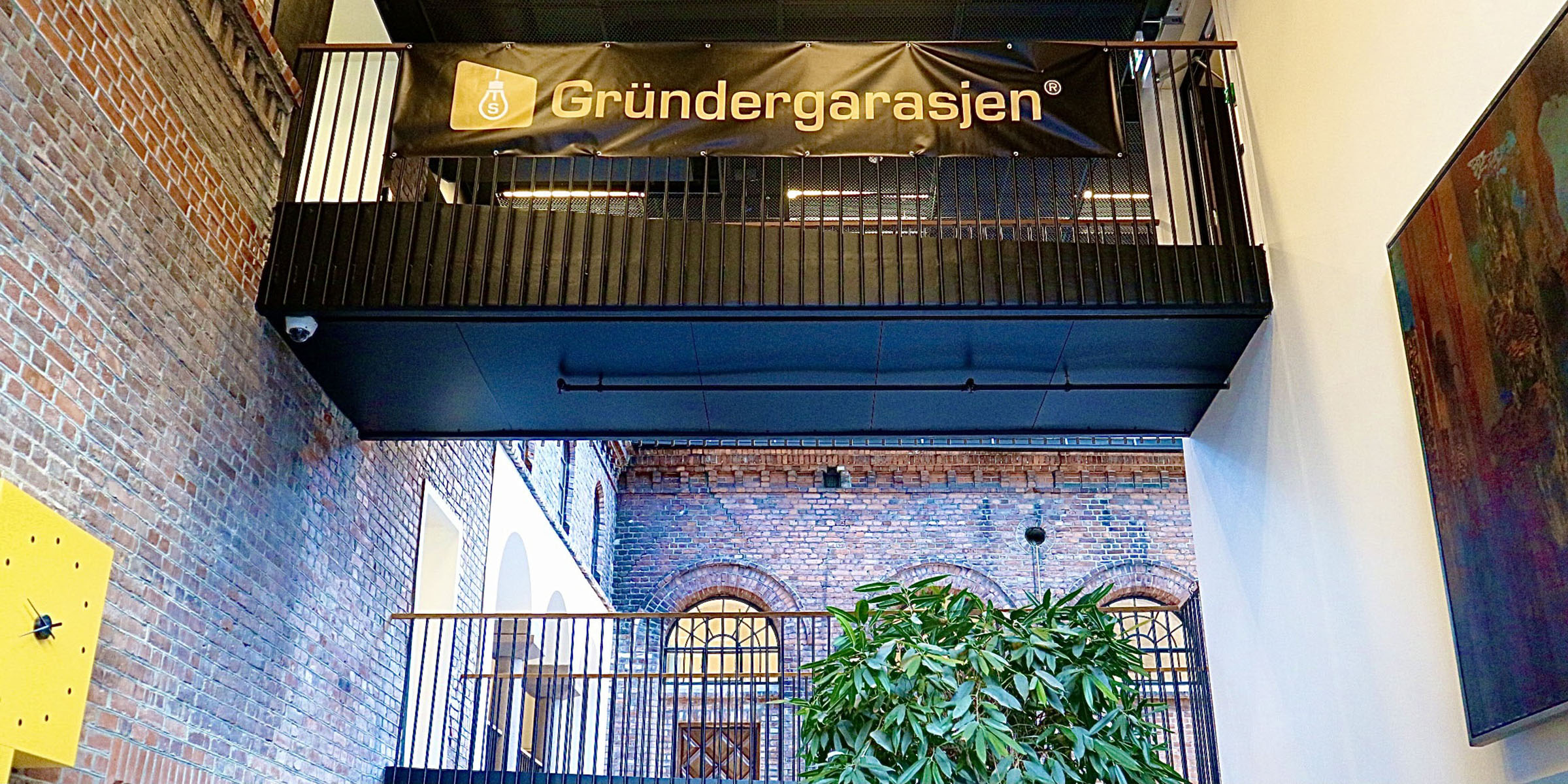 Bildet viser et banner med Gründergarasjen festet på en gangbro øverst i et rom der det er høyt under taket. Det er vegger av murstein til venstre og bak, og nederst kan vi se en plante og en klokke. Store vinduer slipper inn mye lys.