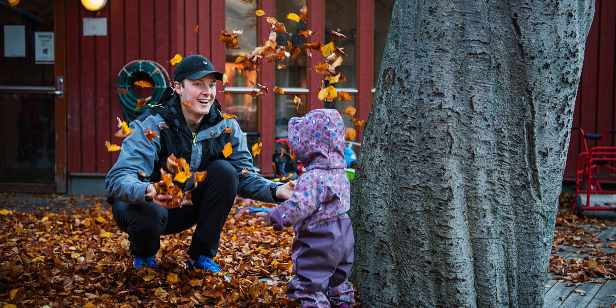 Foto av ung mann som er ute og kaster høstløv opp i luften, foran et lite barn i parkdress.