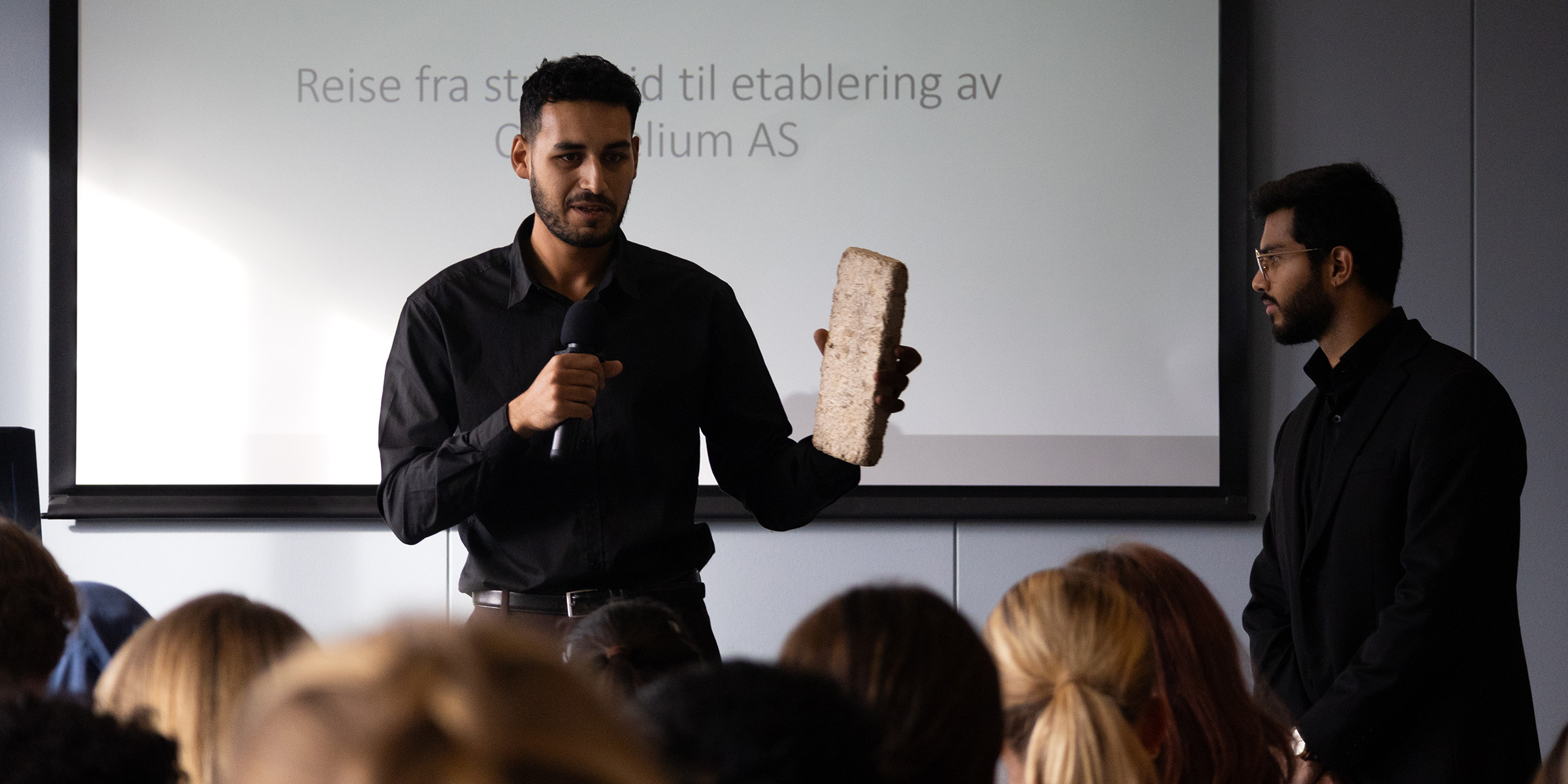 En mann med mikrofon i hånda holder opp en brikke laget av sopp for publikum. En annen mann står vendt sidelengs ved siden av. 