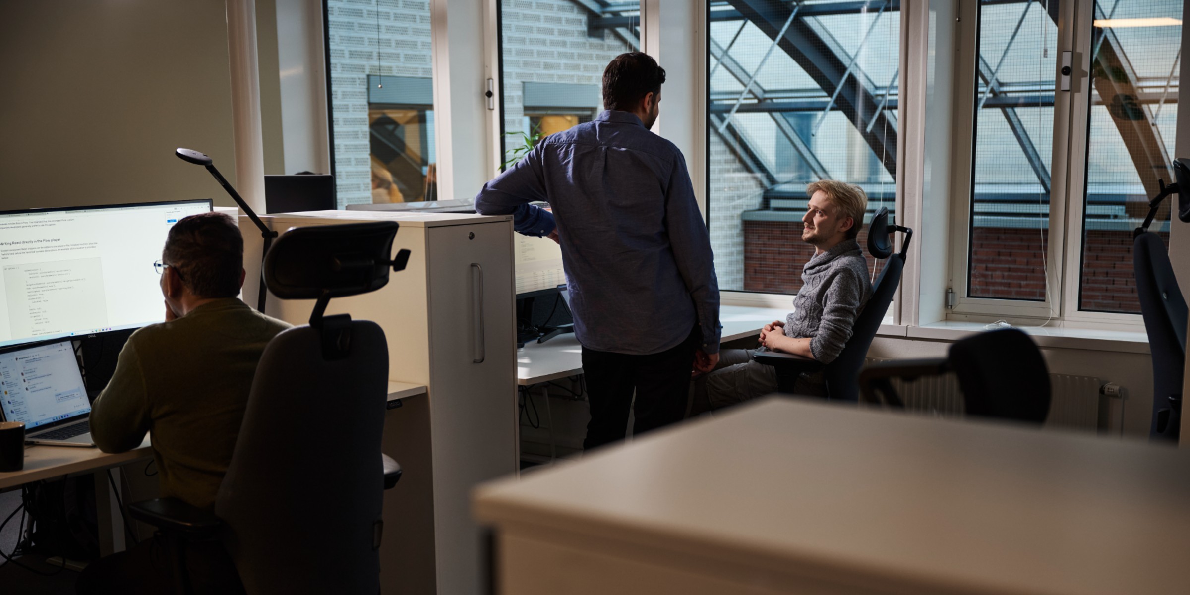 Bilde av flere personer inne på et kontor. To stykker sitter foran PC-en.