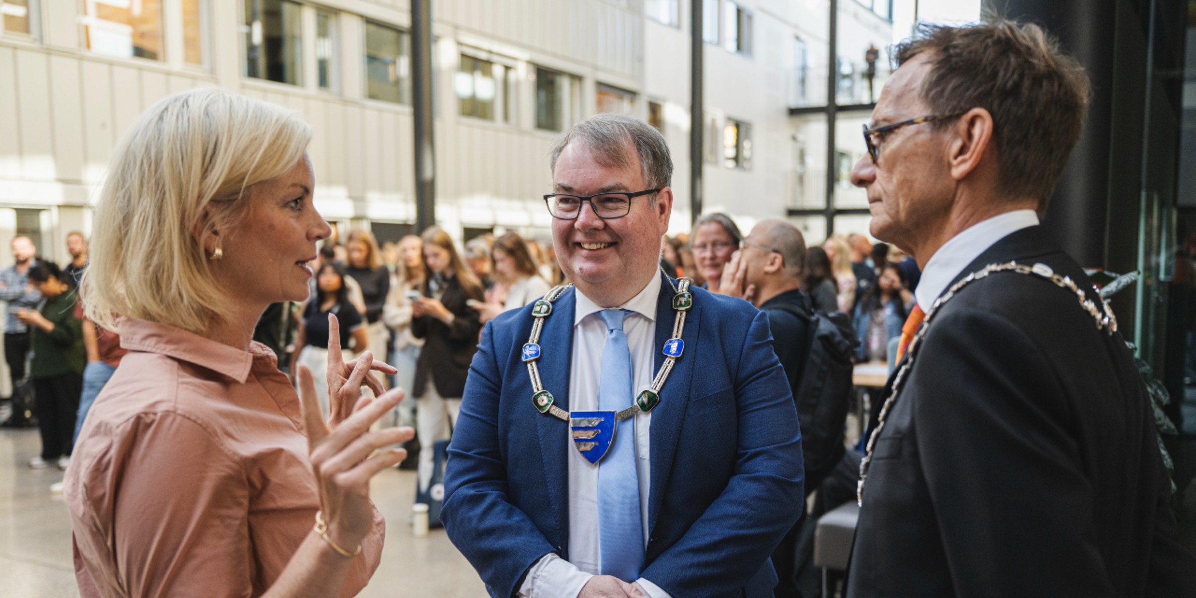 Foto av statsråd Sigrun Aasland, Lillestrøm-ordfører Kjartan Berland og rektor Christen Krogh som snakker sammen med mange studenter stående i bakgrunnen inne på campus Kjeller.