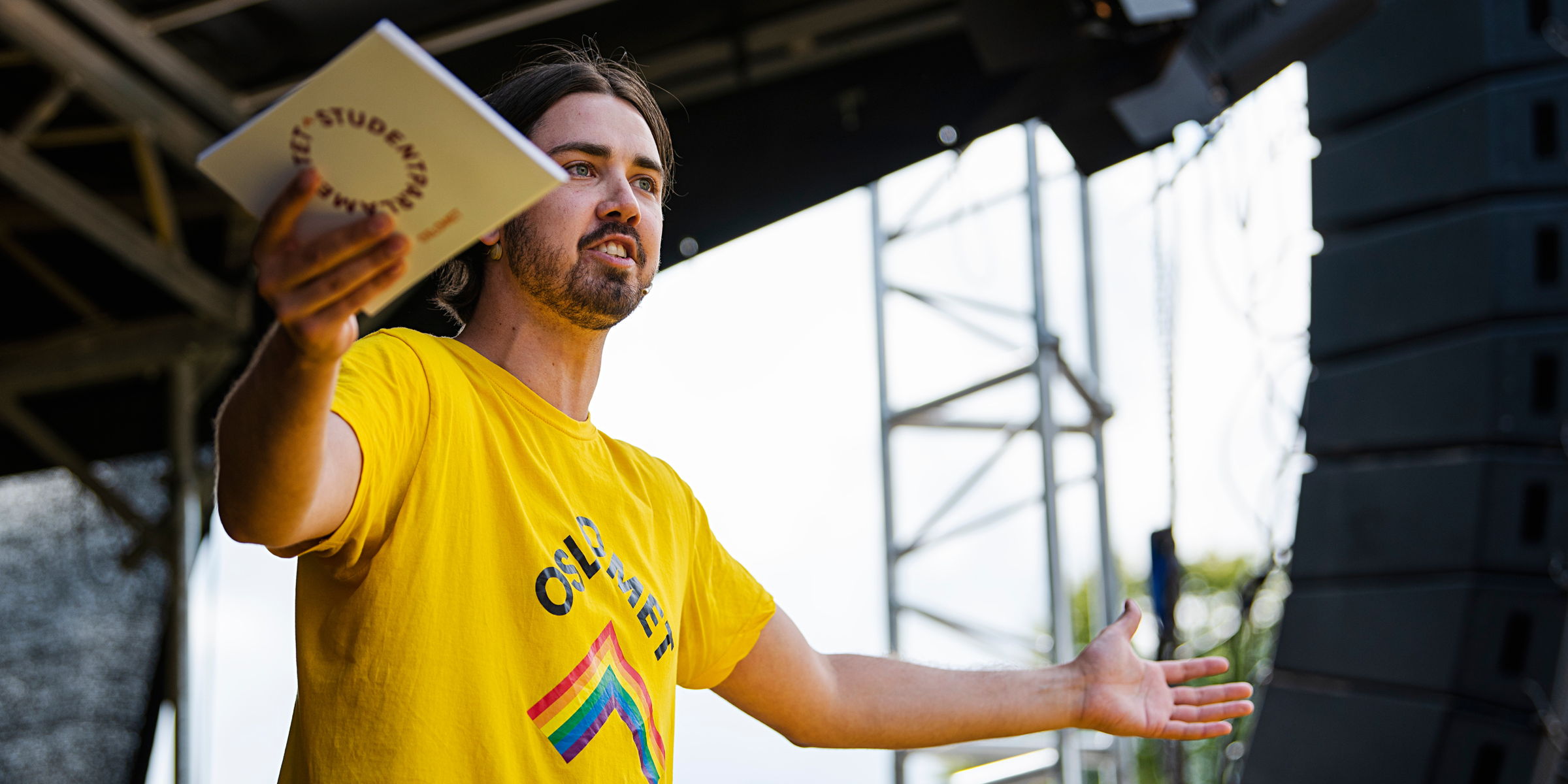 Student in yellow OsloMet t-shirt speaks on stage with open arms. He holds a Student Parliament speech card in one hand.