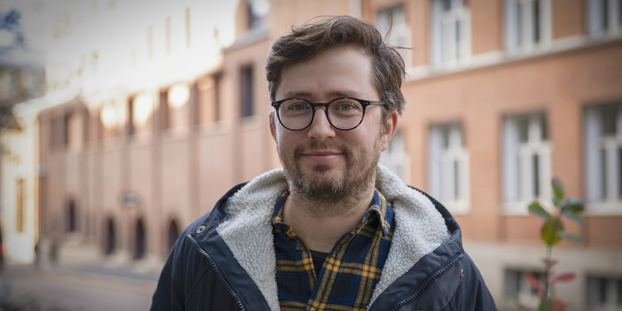 Colour portrait of a man in a blue jacket. In the backgroud there is a brick building.