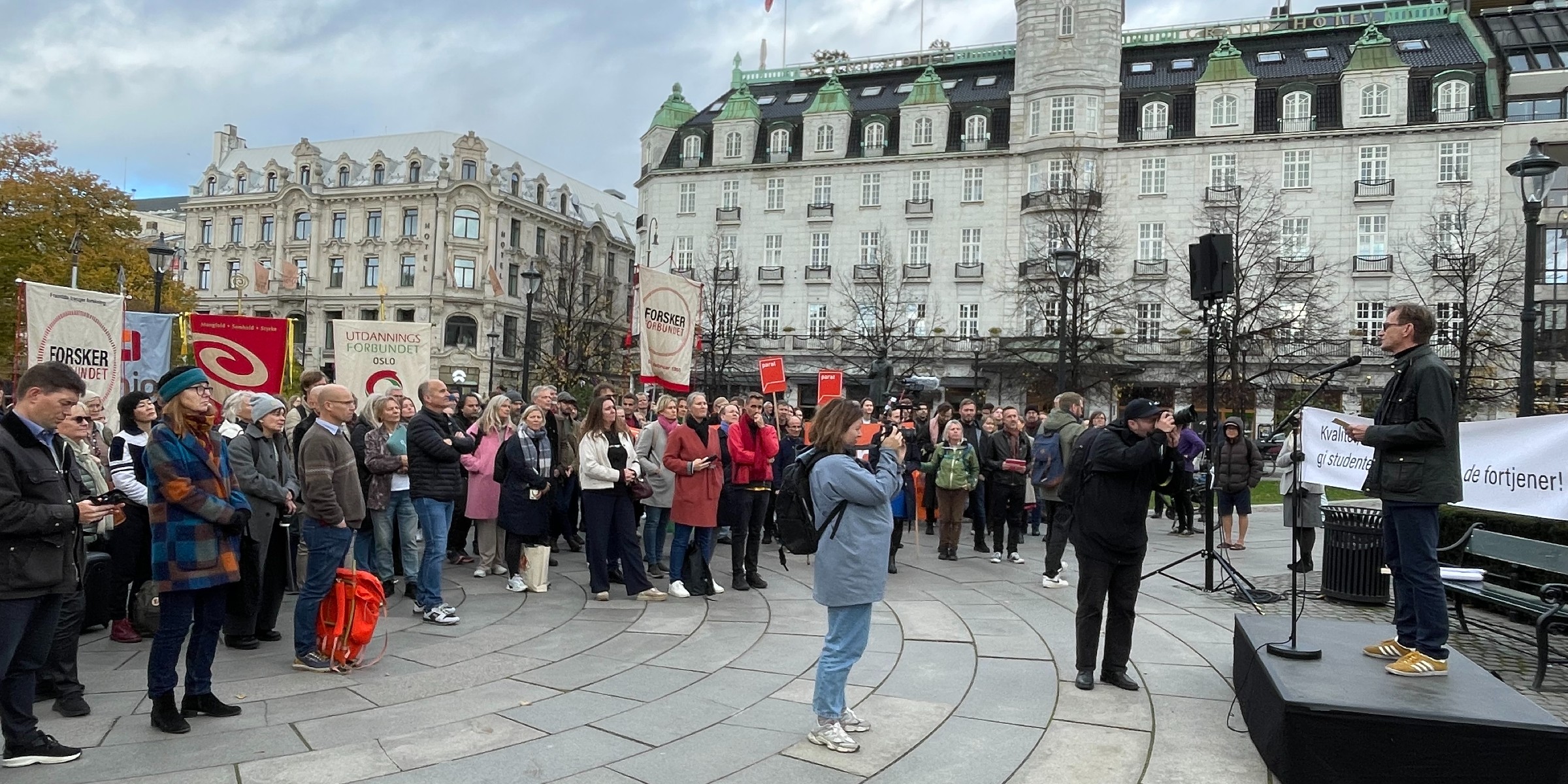 Foto av en samling demonstranter, flere med faner, stående på en plass utenfor Stortinget i Oslo sentrum. Rektor holder appell på talerstol.