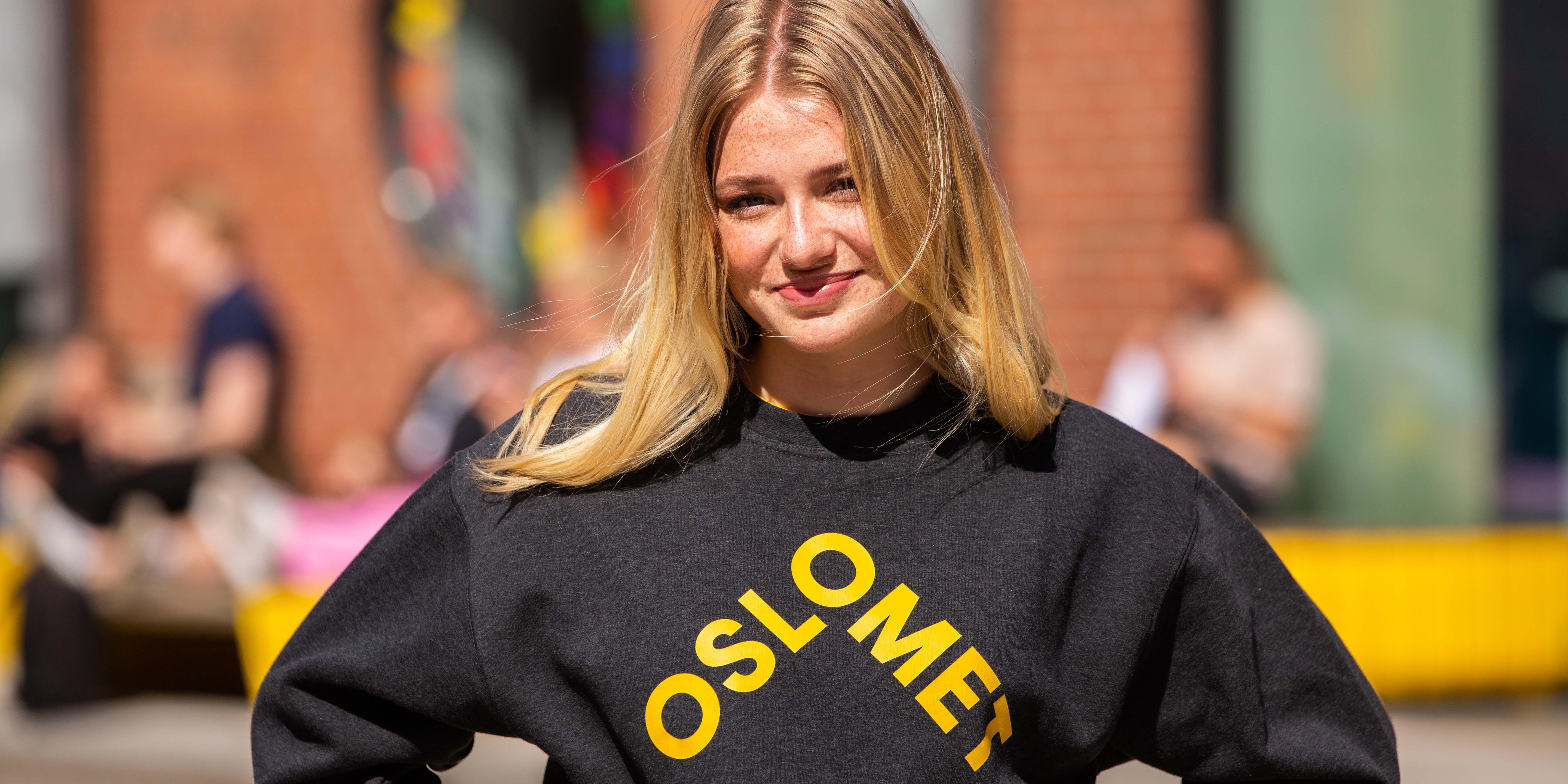 A student with blond hair wearing a grey college jumper with a big yellow OsloMet logo in the front