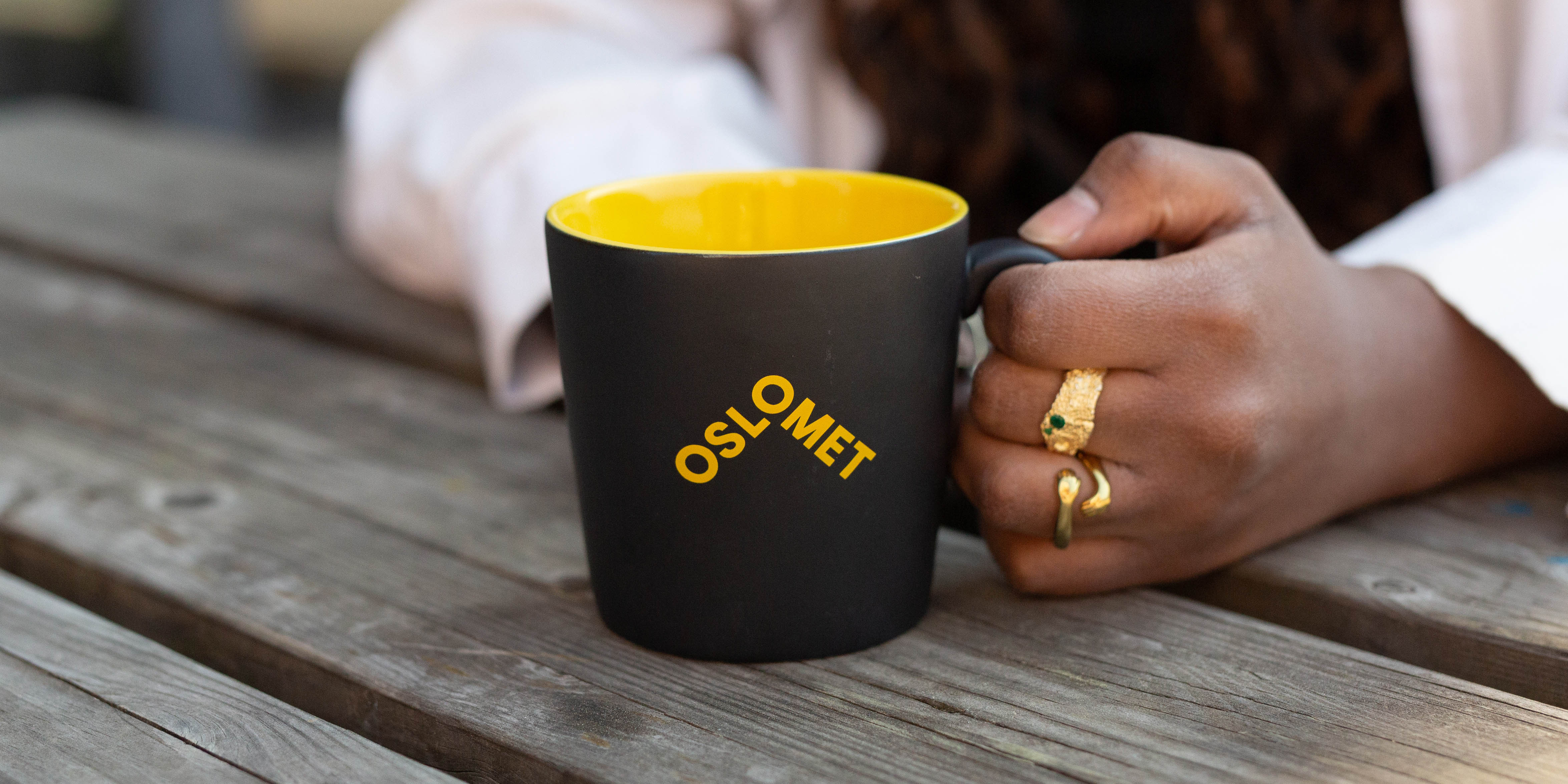 En svart kaffekopp. Kaffekoppen er gul på innsiden, og har en gul OsloMet-logo på forsiden