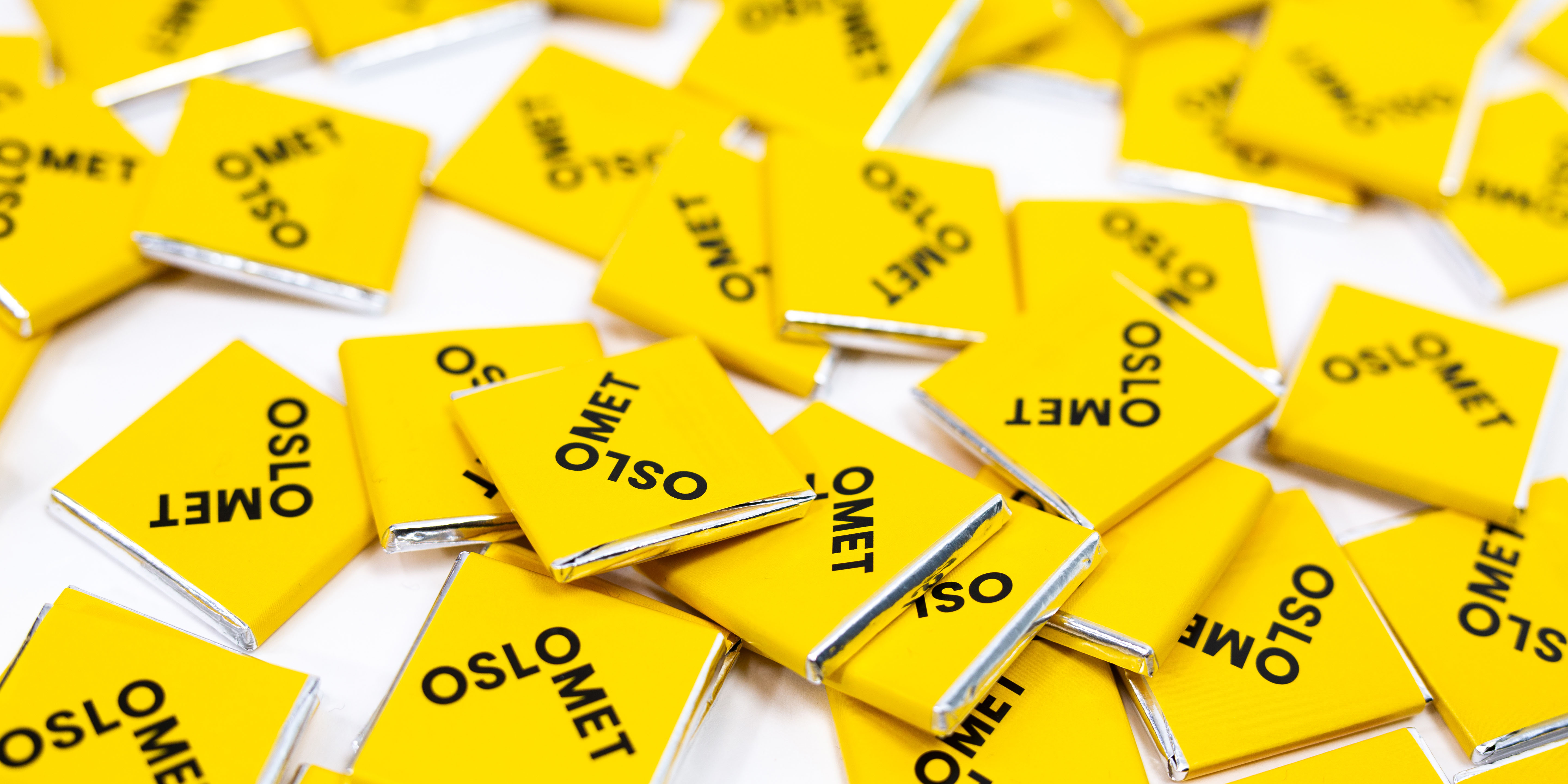 A lot of chocolates spred out on a white surface. The chocolates are individually wrapped in yellow paper, and have a black logo in front