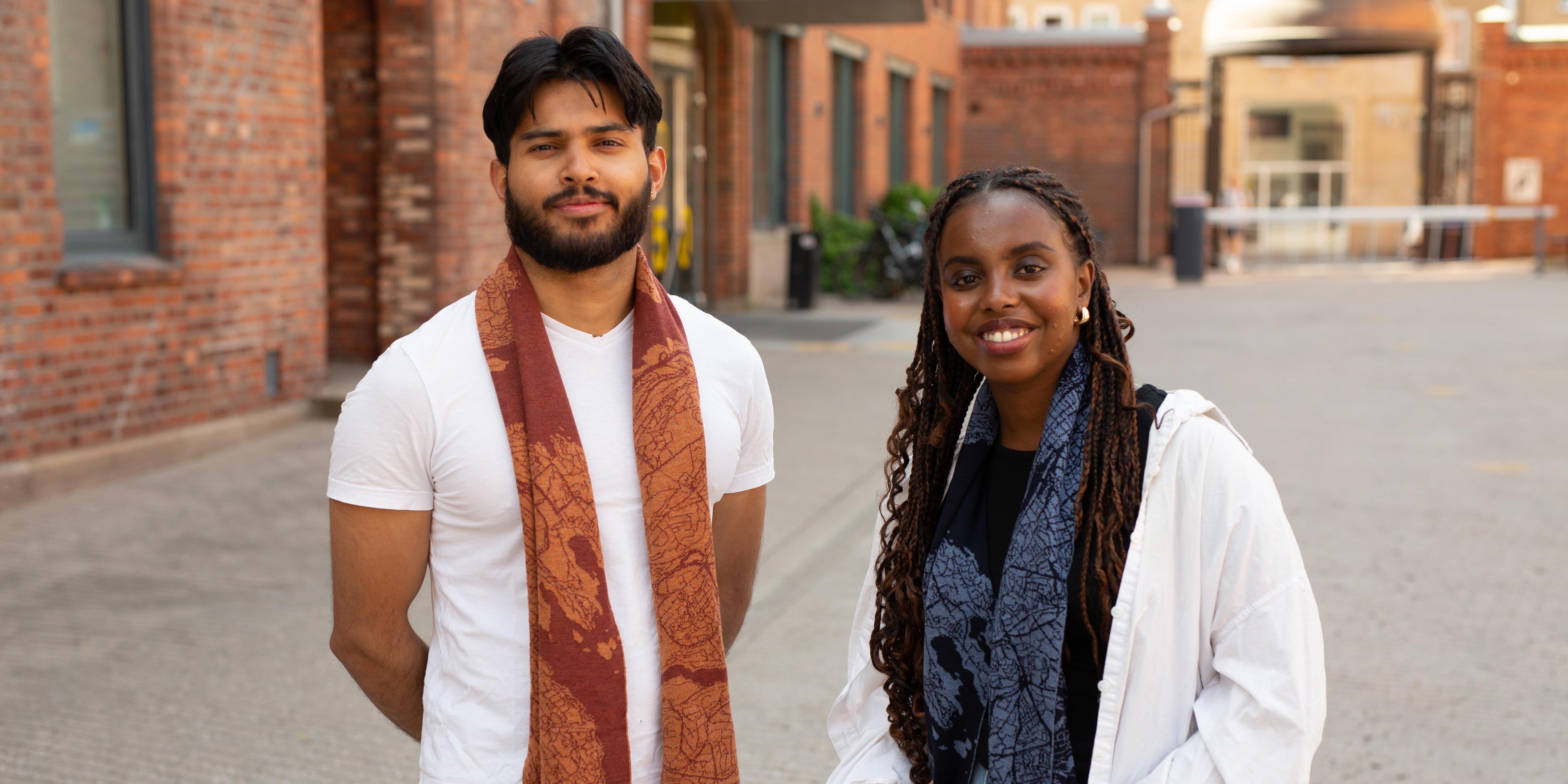 Two students on campus. The boy has a red scarf around his nexk, and the girl has a similar scarf, but in blue