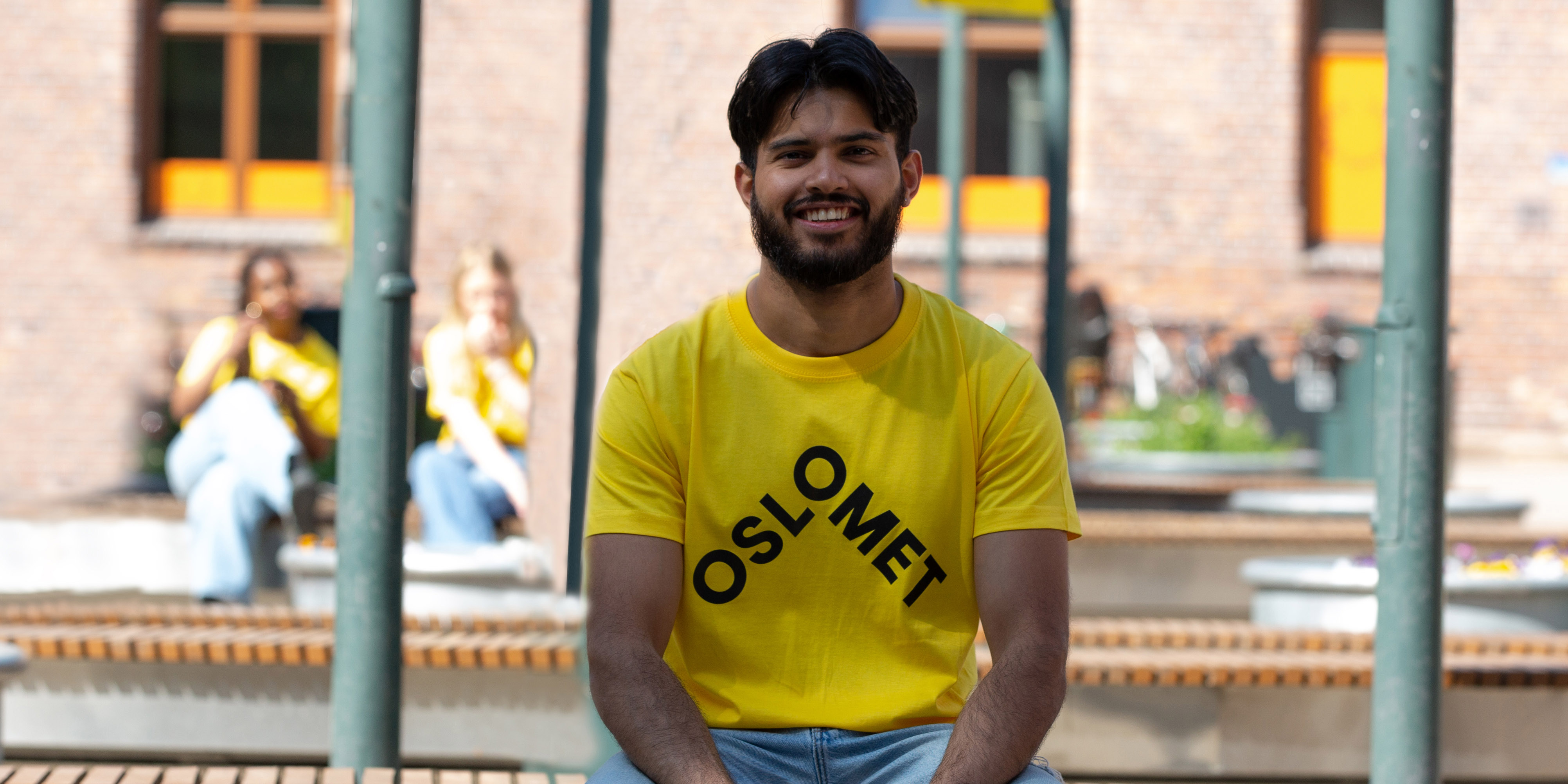 A boy is sitting on a bench. He is wearing a yellow t-shirt with the OsloMet-logo in front