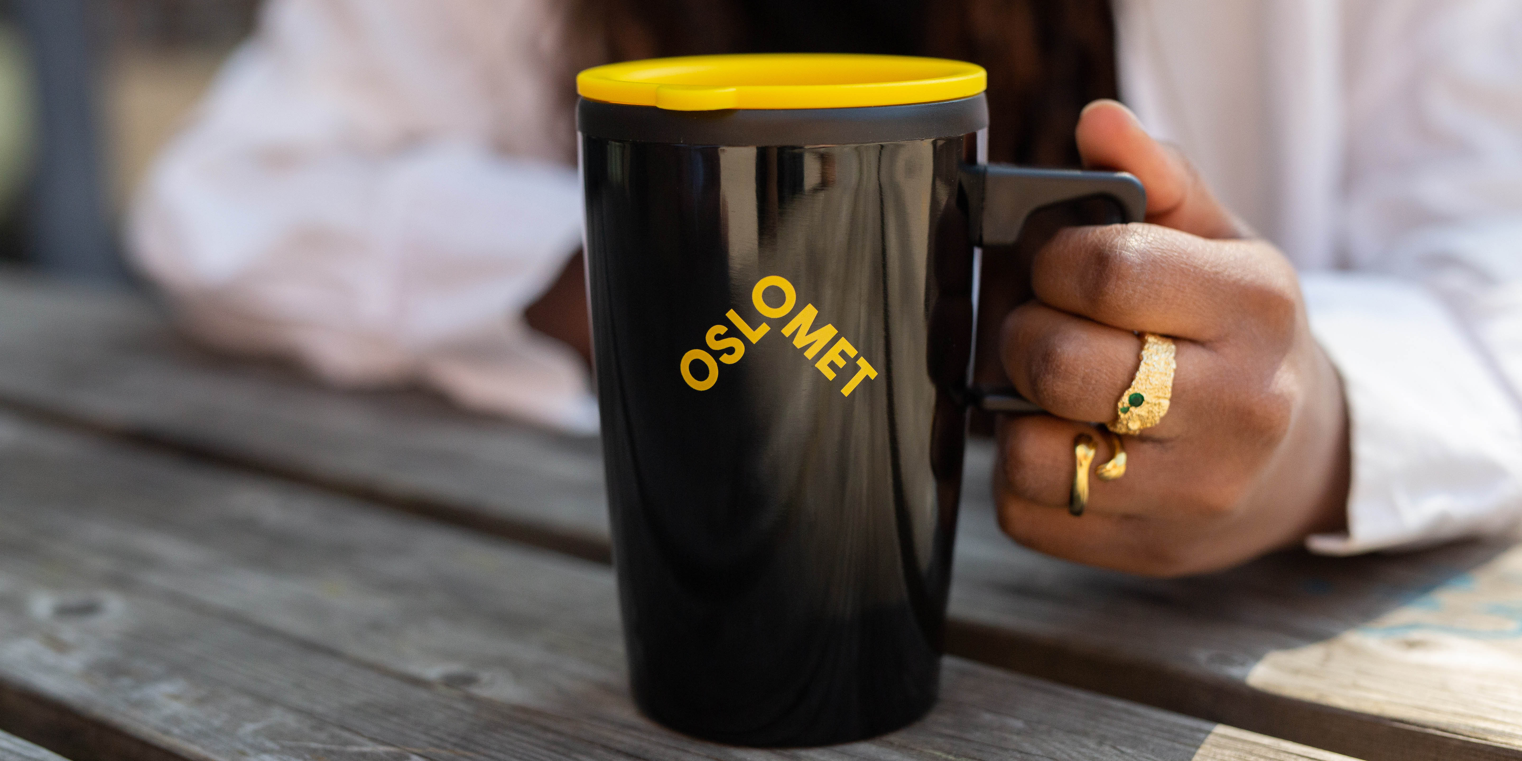 A black cup with a yellow OsloMet logo. The cup has a yellow lid